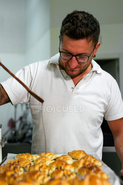 Homem focado padeiro em óculos e roupas casuais de pé e derramando xarope doce em croissants frescos assados na cozinha — Fotografia de Stock