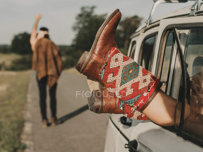 Crop hippie fêmea anônimo em botas ornamentais deitado em automóvel velho temporizador estacionado na estrada perto de homem sem rosto durante a viagem na natureza — Fotografia de Stock
