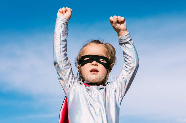 Снизу маленькая девочка в костюме супергероя поднимает протянутые кулаки за демонстрацию силы, стоя на фоне голубого ясного неба — стоковое фото