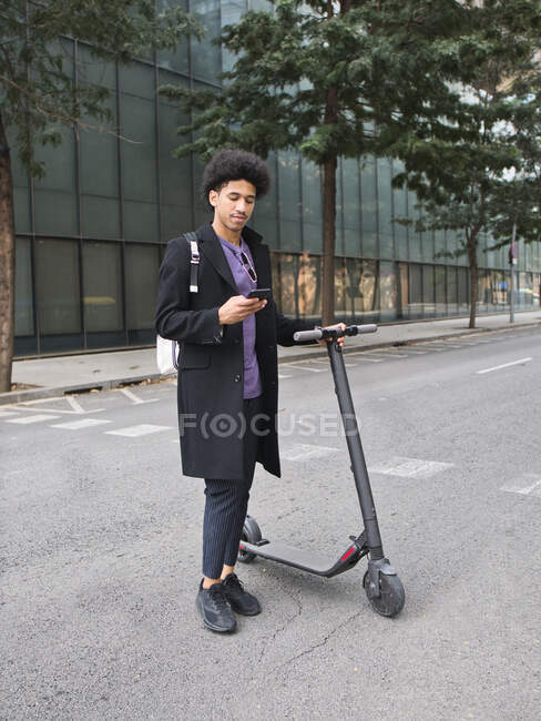 Full body of confident young African American male millennial with dark curly hair in stylish outfit messaging on smartphone while standing on city street with electric scooter — Stock Photo