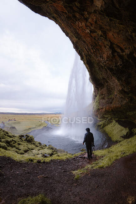Rückenansicht eines anonymen männlichen Reisenden in warmem Outfit, der in einer Felshöhle steht und den malerischen schnellen Seljalandsfoss Wasserfall unter wolkenverhangenem Himmel in Island bewundert — Stockfoto