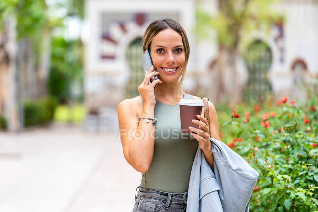 Cheerful female with takeaway coffee having phone conversation and looking at camera while standing on street near green bushes on blurred background — Stock Photo