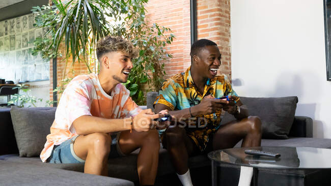 Amigos alegres multirraciales con mandos en las manos sentados en el sofá mientras juegan videojuegos juntos en la sala de estar ligera con planta verde - foto de stock