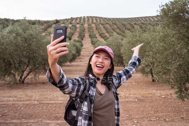 Deliziosa viaggiatrice asiatica a braccia aperte che si autoritrae sullo smartphone mentre si trova in piantagione con ulivi in campagna — Foto stock