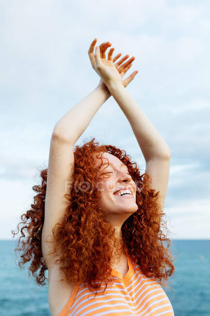 Восхитительная женщина с кудрявыми длинными рыжими волосами в трикотажном свитере стоит с поднятыми руками и закрытыми глазами на фоне синего моря — стоковое фото