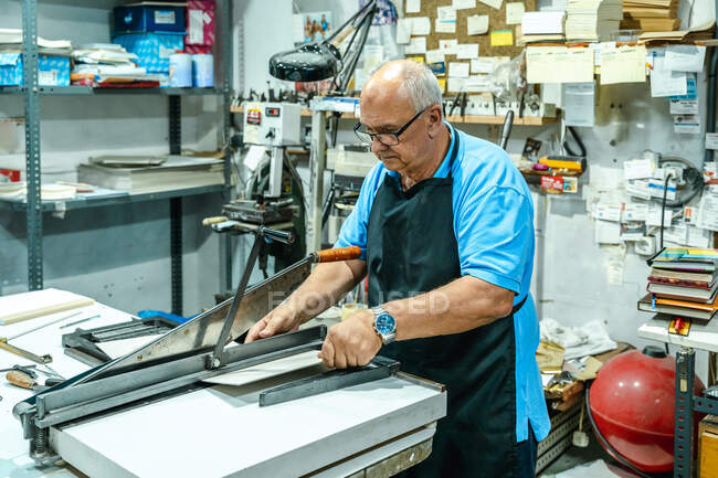Focused elderly male artisan in apron and eyeglasses cutting carton piece with machine while working in printing house — Stock Photo