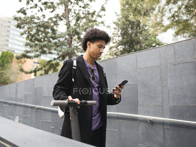 Confident young African American male millennial with dark curly hair in stylish outfit messaging on smartphone while walking on city street with electric scooter — Stock Photo