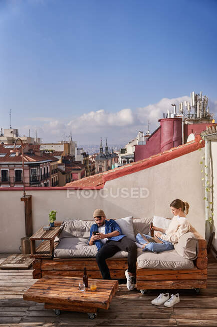 Высокий угол урожая молодая женщина сидит со скрещенными ногами и с помощью нетбука возле мужского соседа прокрутки сотового телефона — стоковое фото