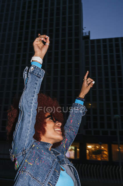 Mujer feliz con peinado afro en traje elegante de pie con los brazos levantados en la calle con edificios residenciales en el tiempo de la noche - foto de stock