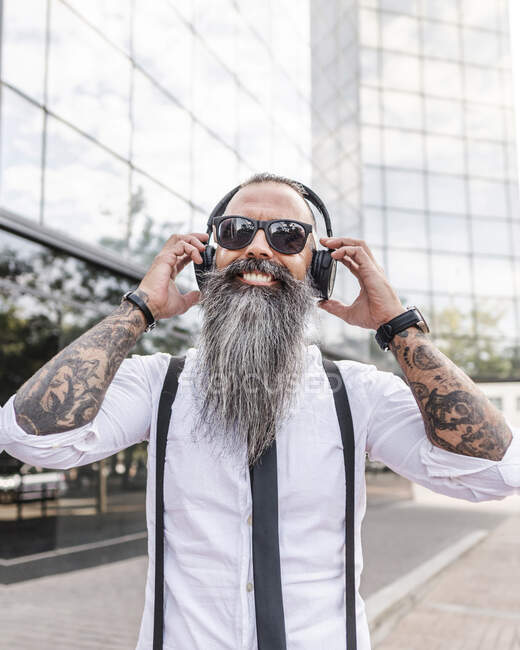 Happy bearded hipster male in formal wear and sunglasses listening to music with wireless headphones while standing near modern buildings — Stock Photo
