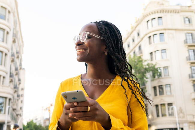 Mujer afro-americana positiva en anteojos mensajería de texto en el teléfono celular mientras está de pie en la calle con edificios residenciales en la calle en la ciudad - foto de stock
