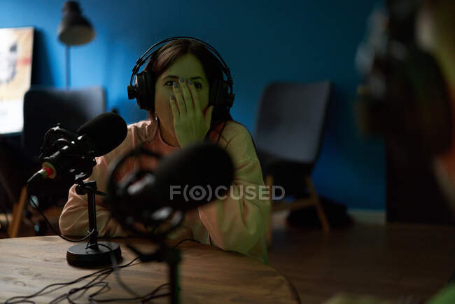 Jeune animatrice radio en vêtements décontractés et écouteurs assis à table avec microphone et communiquant avec un collègue anonyme lors de l'enregistrement de podcast en studio — Photo de stock