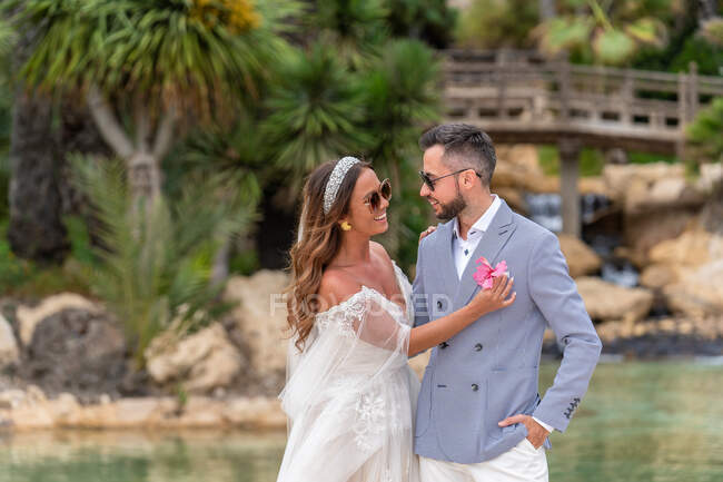 Smiling married couple in wedding clothes and sunglasses standing near lake and green trees with plants while looking at each other and woman giving man flower in daytime in park — Stock Photo
