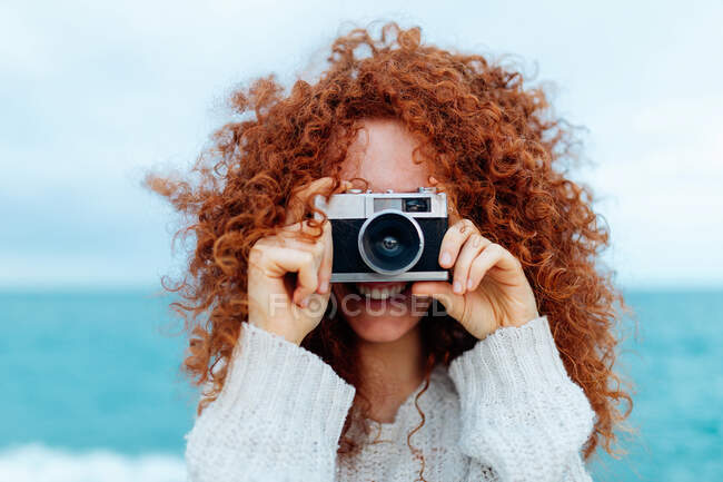 Mujer pelirroja positiva en suéter de punto tomando fotos en cámara fotográfica retro en la costa del mar - foto de stock