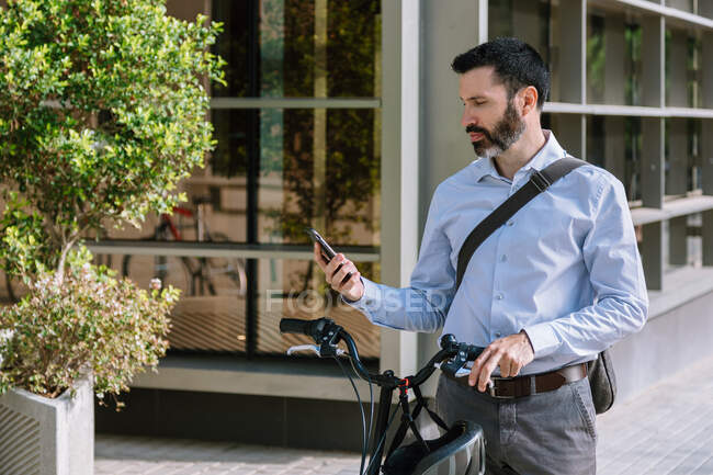 Чоловік менеджер ходить з велосипедом і читає повідомлення на смартфоні в парку — стокове фото