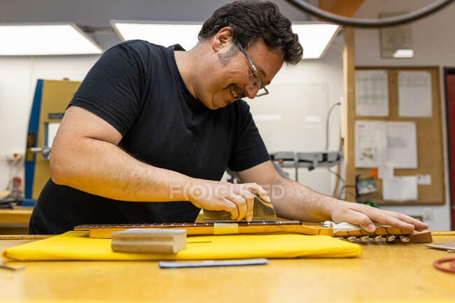 Vue latérale de l'artisan joyeux debout à la table et le manche de broyage de la guitare dans l'atelier professionnel — Photo de stock