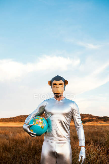 Homem anônimo em terno de látex de prata com máscara de macaco geométrico olhando para a câmera e segurando globo na natureza — Fotografia de Stock