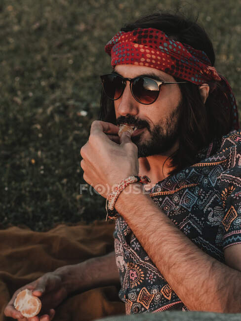 Hippie-Mann mit Sonnenbrille isst Mandarine, während er bei Sonnenuntergang auf einer Wiese in der Natur liegt — Stockfoto