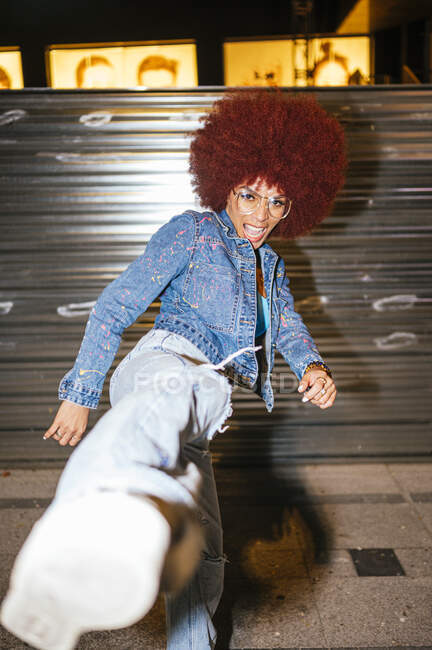 Atractiva hembra con peinado afro en traje de moda pateando el aire y mirando a la cámara mientras está de pie en la calle por la noche - foto de stock