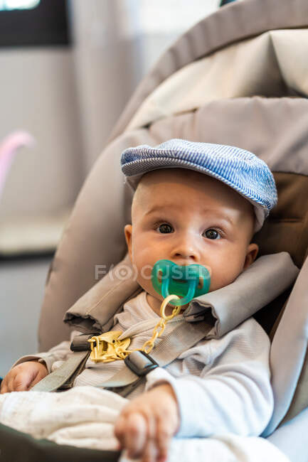 Adorable baby in hat looking away and sucking pacifier while sitting in comfortable stroller with safety harness in light room — Stock Photo