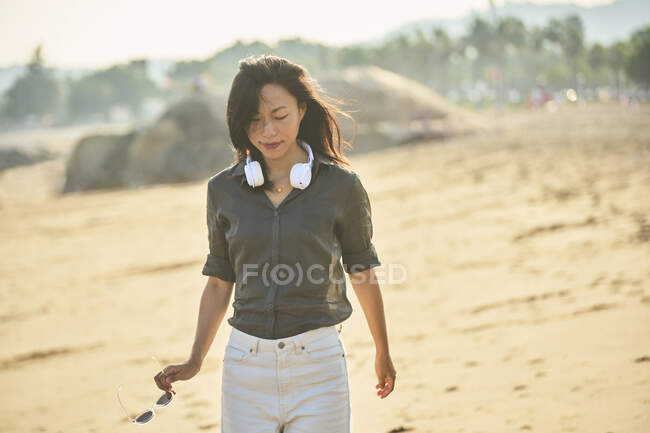 Side view of peaceful thoughtful Asian female looking down listening to song from wireless headphones while walking on sandy shore — Stock Photo