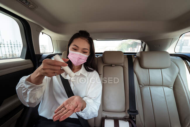 Mulher étnica em máscara derramando desinfetante em mãos enquanto montava no banco de trás no táxi — Fotografia de Stock