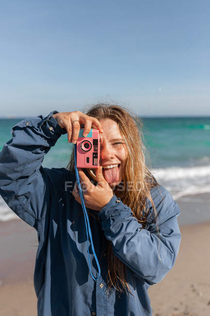 Cheerful young female in wet clothes taking photo on camera while standing with tongue out on sandy beach near waving sea — Stock Photo