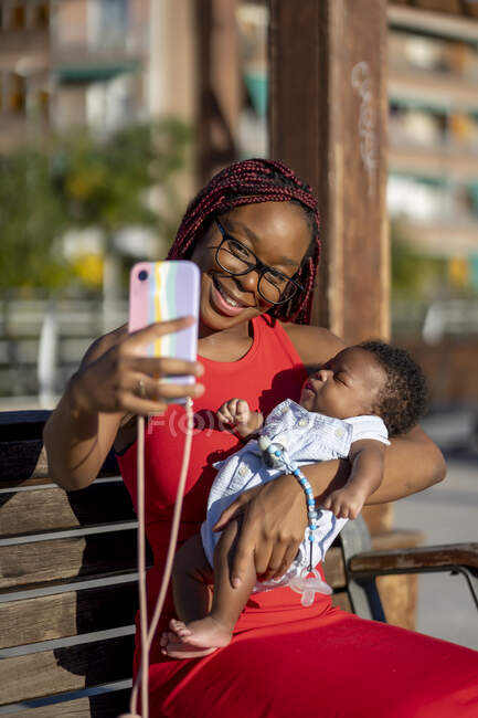 Sonriente madre afroamericana con trenzas en vestido rojo sentado con el bebé a la mano en el banco de madera y tomando selfie en el teléfono inteligente en la calle en el día soleado - foto de stock