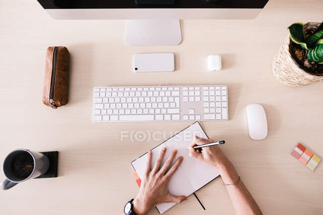 Overhead of crop worker writing in notebook placed on desk with stationery and cup and smartphone near computer and plant - foto de stock