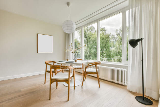 Tavolo rotondo con piatti e vaso con ramoscelli situato vicino alla finestra nella spaziosa sala da pranzo luminosa — Foto stock