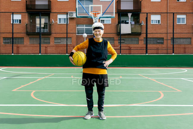 Mujer madura positiva en ropa deportiva y diadema mirando a la cámara mientras está de pie con la pelota durante el juego de baloncesto - foto de stock