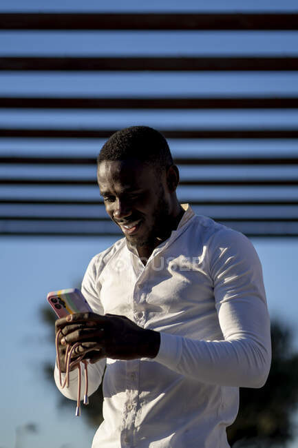Снизу улыбающийся афроамериканец в белой рубашке, стоящий и просматривающий смартфон на улице в солнечный день — стоковое фото