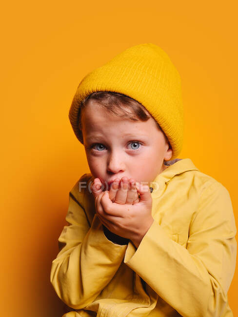 Petit garçon calme avec des yeux bleus dans une veste jaune vif et un chapeau envoyant un baiser d'air et regardant la caméra sur fond jaune en studio — Photo de stock