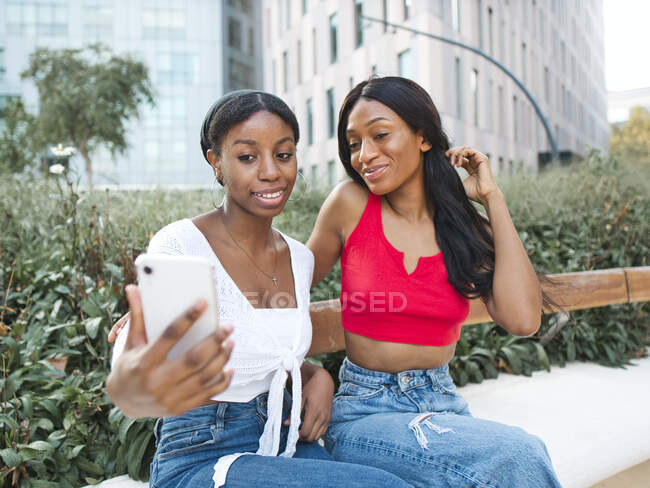 Deliziose amiche afroamericane che si autoritrattano sul cellulare mentre siedono sulla panchina vicino a piante verdi in strada con edifici moderni — Foto stock