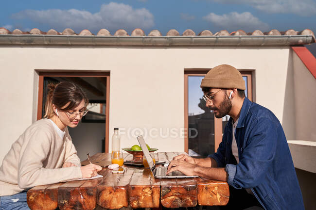 Mujer concentrada en anteojos escribiendo en planificador en la mesa con compañero de piso masculino en auriculares inalámbricos escribiendo en el portátil en el balcón - foto de stock