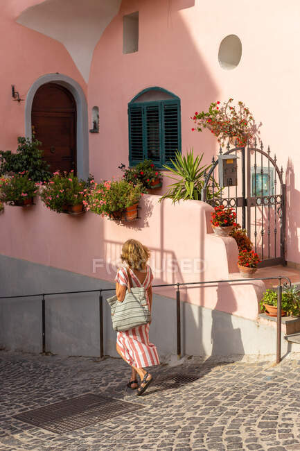 Back view of unrecognizable elegant female tourist in stylish dress walking on sloped narrow street near typical pink residential building during summer holidays on Procida Island — Stock Photo