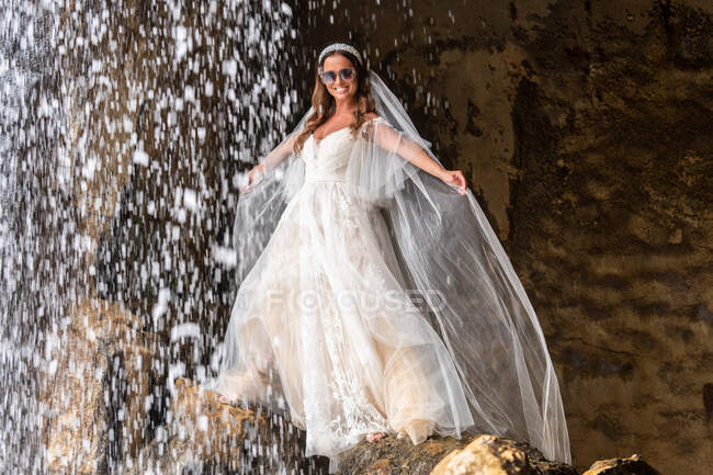 Bride in white wedding dress with veil and sunglasses standing on boulder near falling cascade in nature during holiday celebration on summer day — Stock Photo