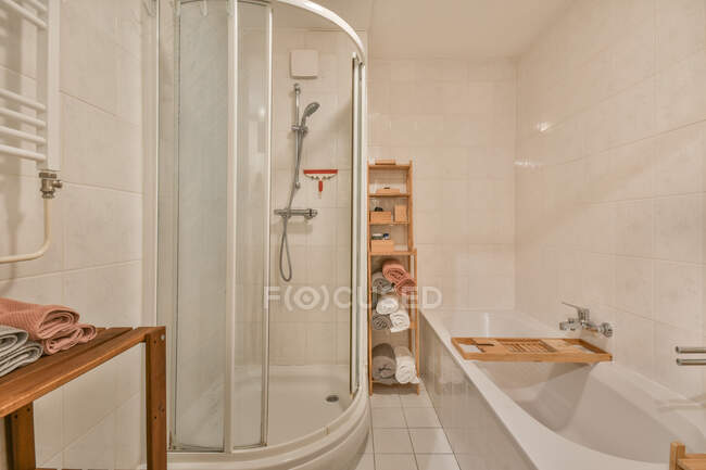 Interior de baño limpio con azulejo claro con bañera blanca y cabina de ducha iluminada con lámparas brillantes y amueblada con estantes de madera equipados con toallas - foto de stock