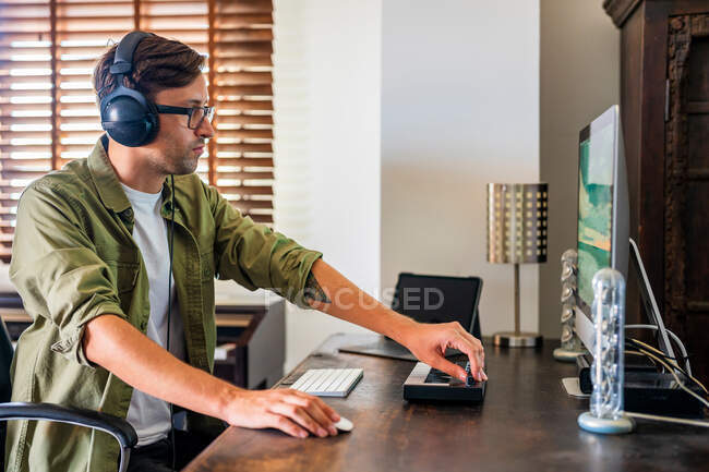 Side view of concentrated guy with headphones sitting at table and using mouse while working on computer — Stock Photo