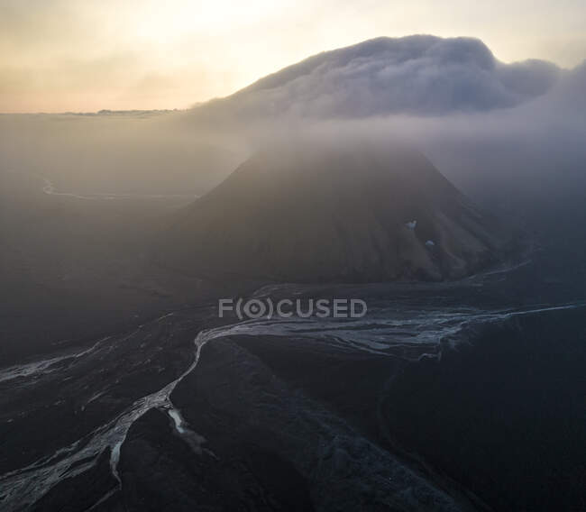 Вулканічна гора з крутою поверхнею, вкритою туманом, розташована поблизу тече річка в дикій природі проти безхмарного неба ввечері — стокове фото