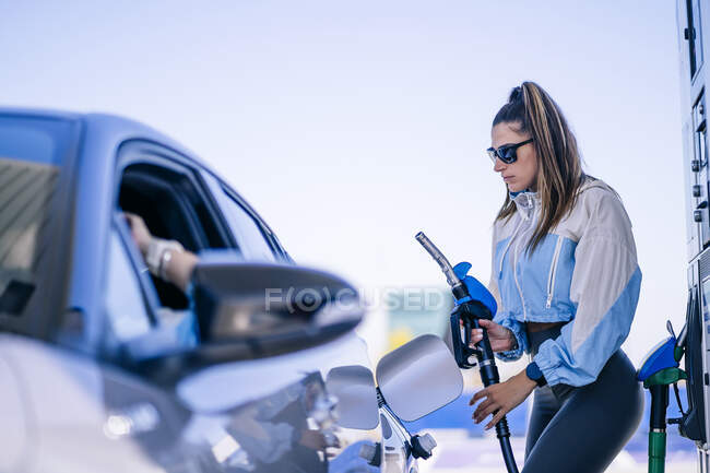 Schlanke Frau mit Sonnenbrille tankt Auto bei Tageslicht mit anonymer Frau — Stockfoto