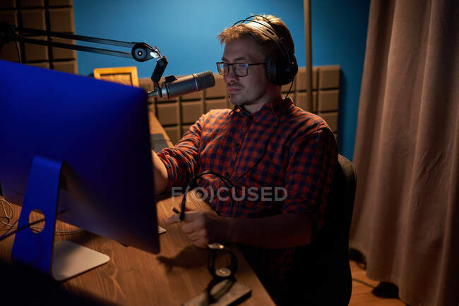 D'en haut jeune homme concentré en chemise à carreaux et lunettes à l'aide d'un ordinateur et parler en micro tout en enregistrant podcast en studio sombre — Photo de stock