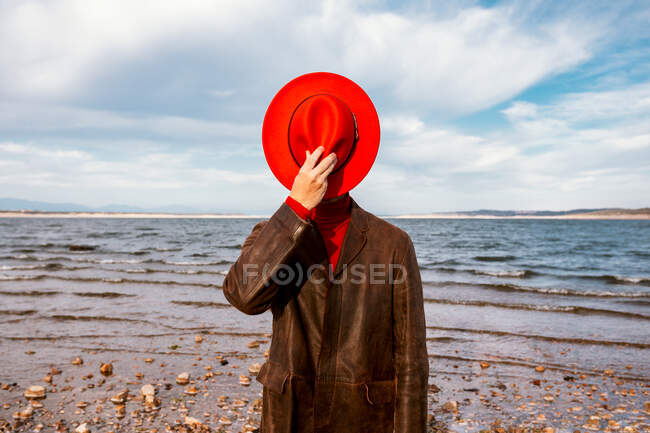 Неузнаваемый человек в красной шляпе и пальто, стоящих на берегу с дроблеными камнями летом — стоковое фото