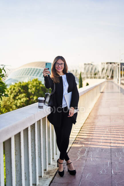 Позитивная женщина в стильном формальном наряде стоящая с ноутбуком и делающая автопортрет на сотовом телефоне рядом с перилами моста — стоковое фото