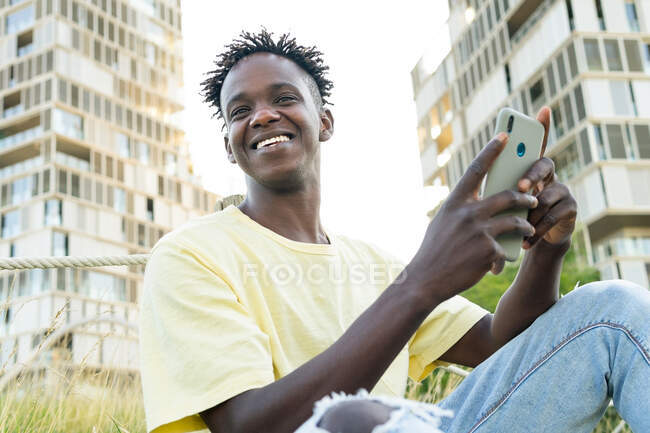 Sonriente joven hombre negro con camiseta de color amarillo claro y pantalones vaqueros azules y zapatillas de deporte sentado en la hierba y el teléfono inteligente de navegación - foto de stock