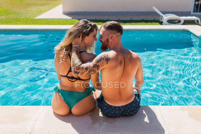 Visão traseira de casal amoroso em trajes de banho olhando uns para os outros enquanto acaricia na borda da piscina em resort tropical — Fotografia de Stock