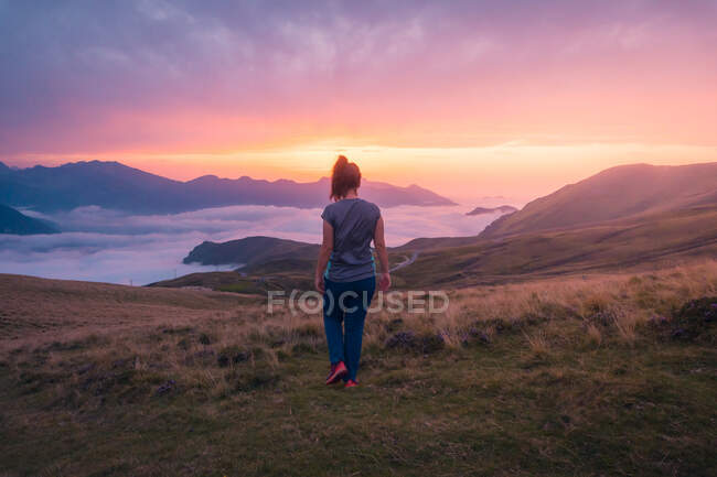 Vista trasera de cuerpo completo de una excursionista femenina irreconocible que camina sobre un campo cubierto de hierba cerca de una espesa niebla blanca contra la cresta de la montaña por la noche - foto de stock