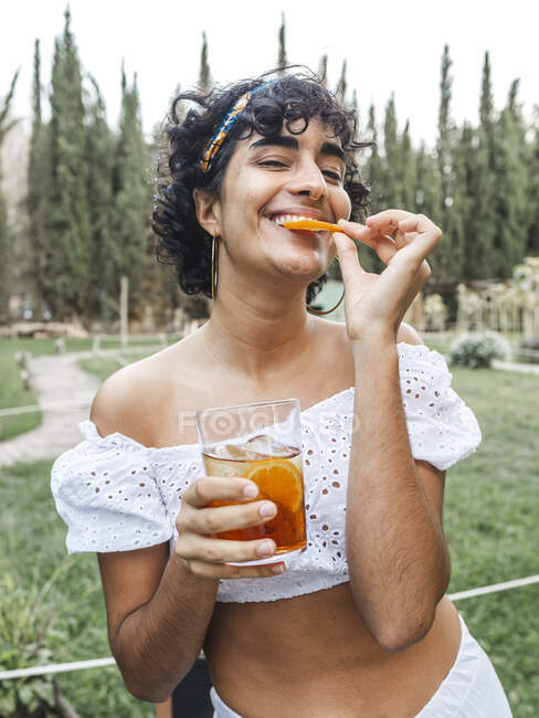 Content ethnic female eating slice of orange while standing with glass of cold cocktail in summer park and looking at camera — Stock Photo
