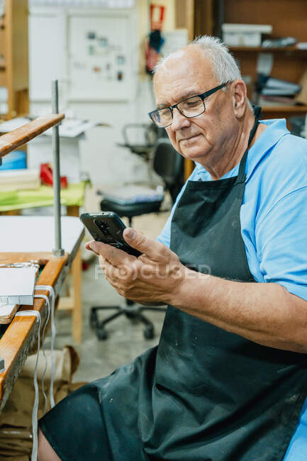 Concentrated senior male master in apron and eyeglasses using mobile phone while sitting at workbench during printing process in studio — Stock Photo
