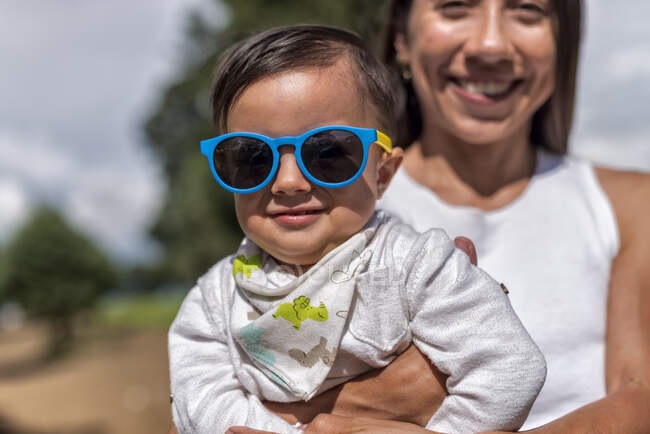 Положительная мать, держащая симпатичного ребенка в солнцезащитных очках, стоя в сельской местности с высокими зелеными деревьями на размытом фоне — стоковое фото